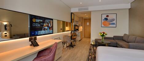 Una habitación de hotel moderna con una cama grande, un escritorio con un teléfono, una zona de estar con sofá y sillones, una pequeña mesa con un jarrón de flores y un espejo grande que refleja la habitación.