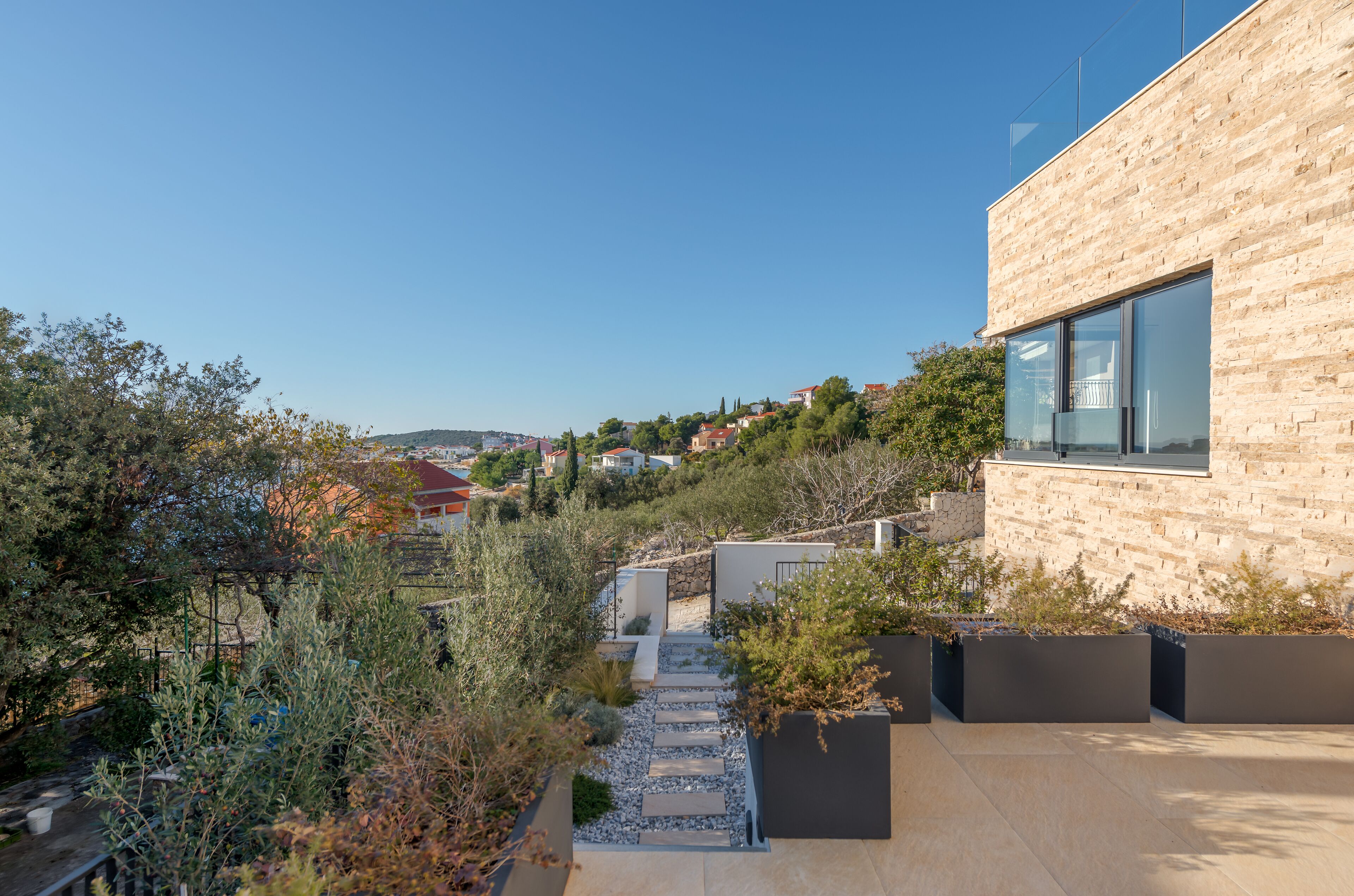 A modern building with a stone facade and large windows, surrounded by greenery and a clear sky.
