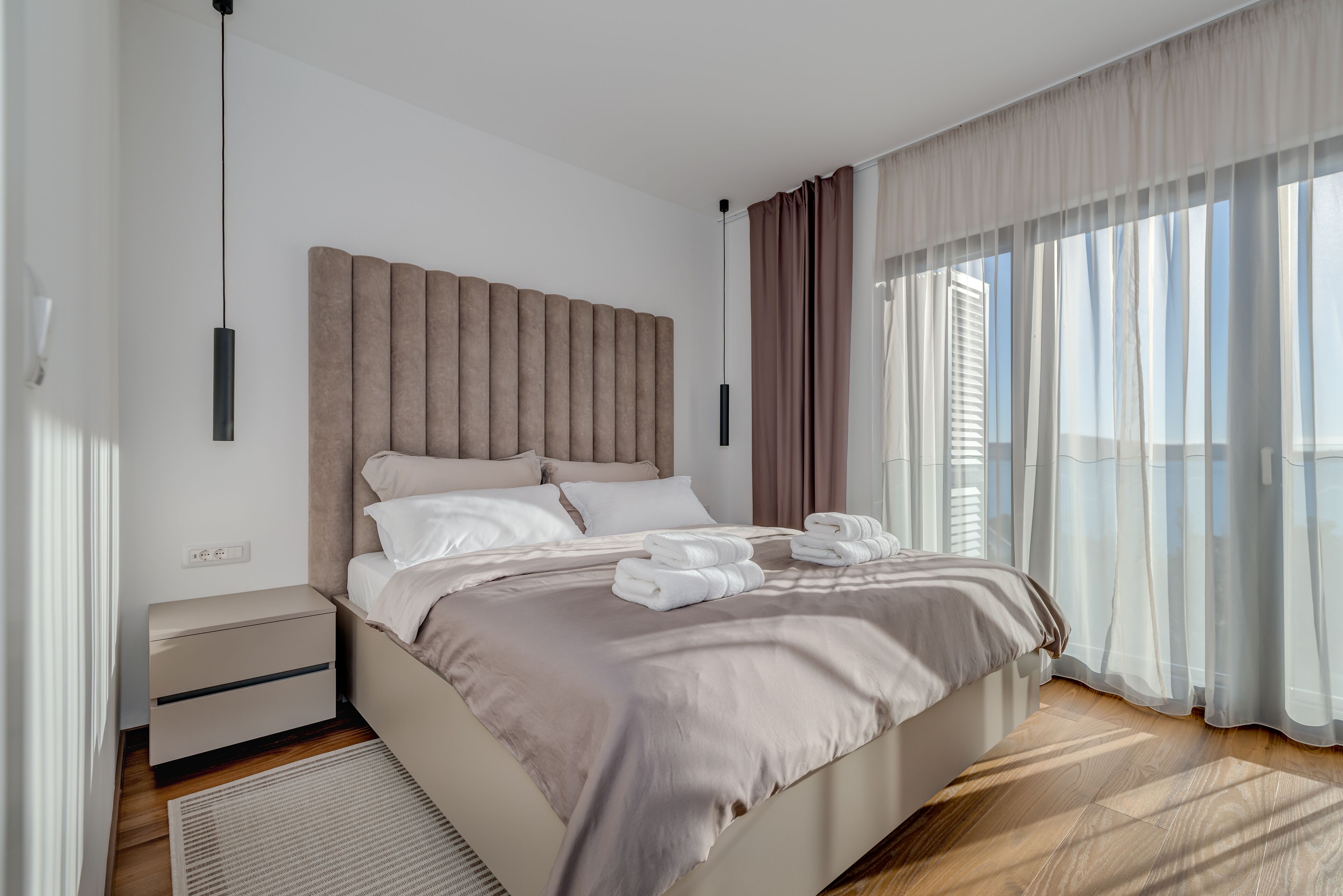 A modern bedroom with a large bed, a nightstand, and a view of the city through sheer curtains.