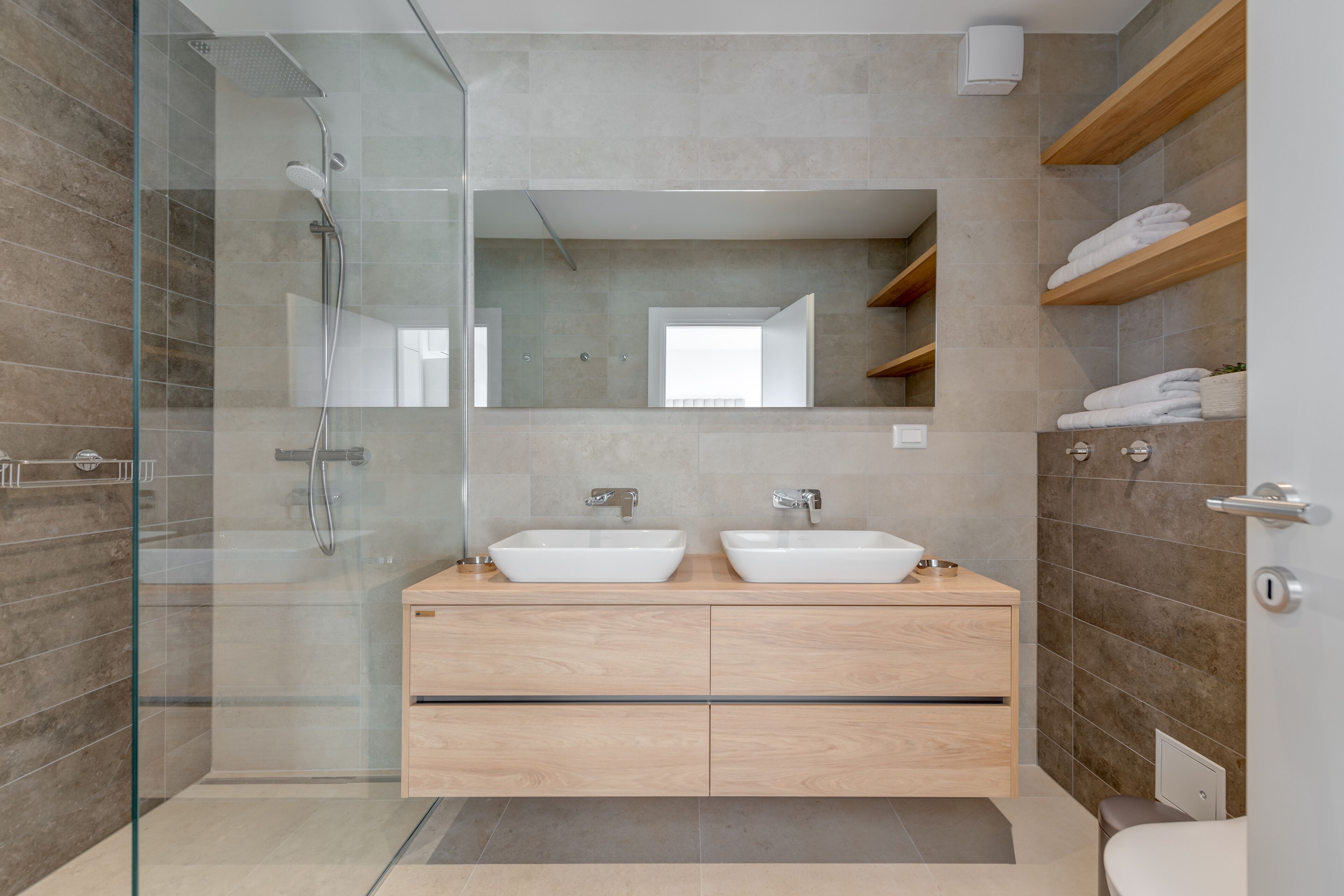 A modern bathroom with a glass shower enclosure, two sinks, wooden vanity, and built-in shelves with towels.