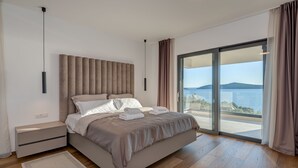 A modern bedroom with a large bed, a nightstand, a bedside table, and a view of the sea through a sliding glass door. - Sea Bliss Luxury Villa - Sevid (Marina)