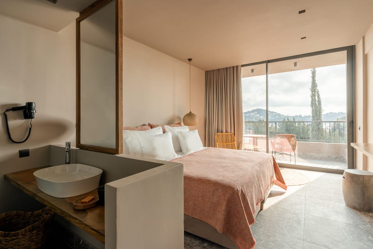 A modern hotel room with a large bed, a round sink, a mirror, and a balcony with a view of the hills.