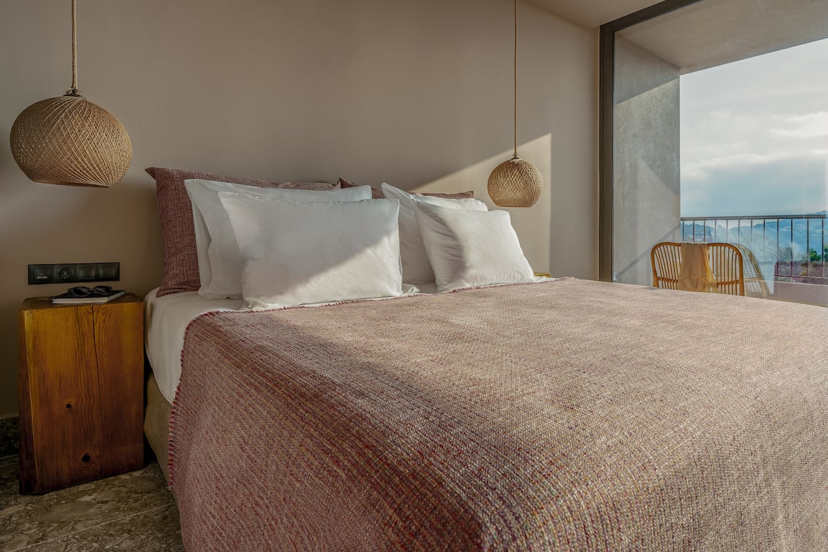 A modern bedroom with a large bed, two hanging wicker lamps, a wooden nightstand, and a view of the outdoors.