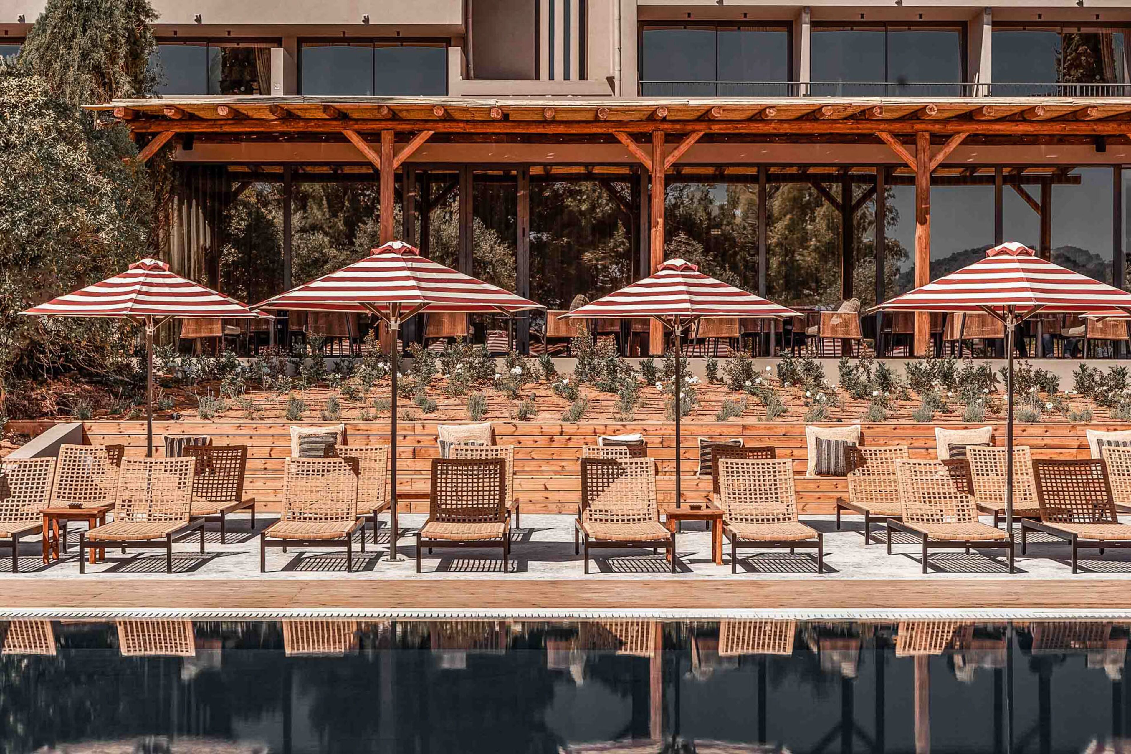 Un área junto a la piscina con sillones de descanso y sombrillas a rayas, una pérgola de madera y un edificio con grandes ventanales al fondo.