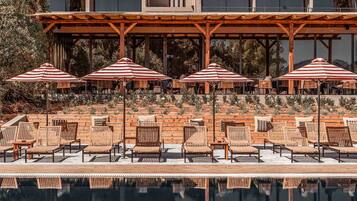 Un área junto a la piscina con sillones de descanso y sombrillas a rayas, una pérgola de madera y un edificio con grandes ventanales al fondo.