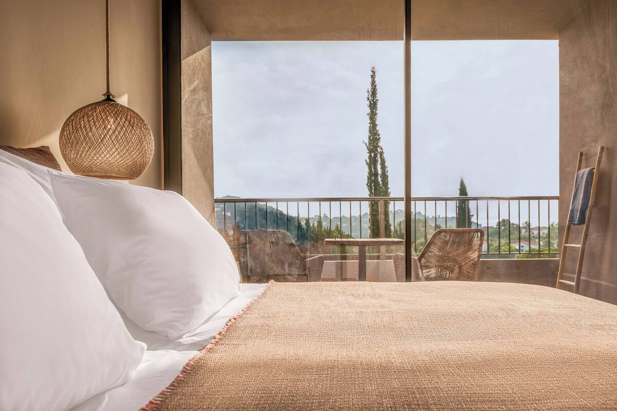 A bedroom with a large bed, a hanging light fixture, and a balcony with a view of trees and hills.