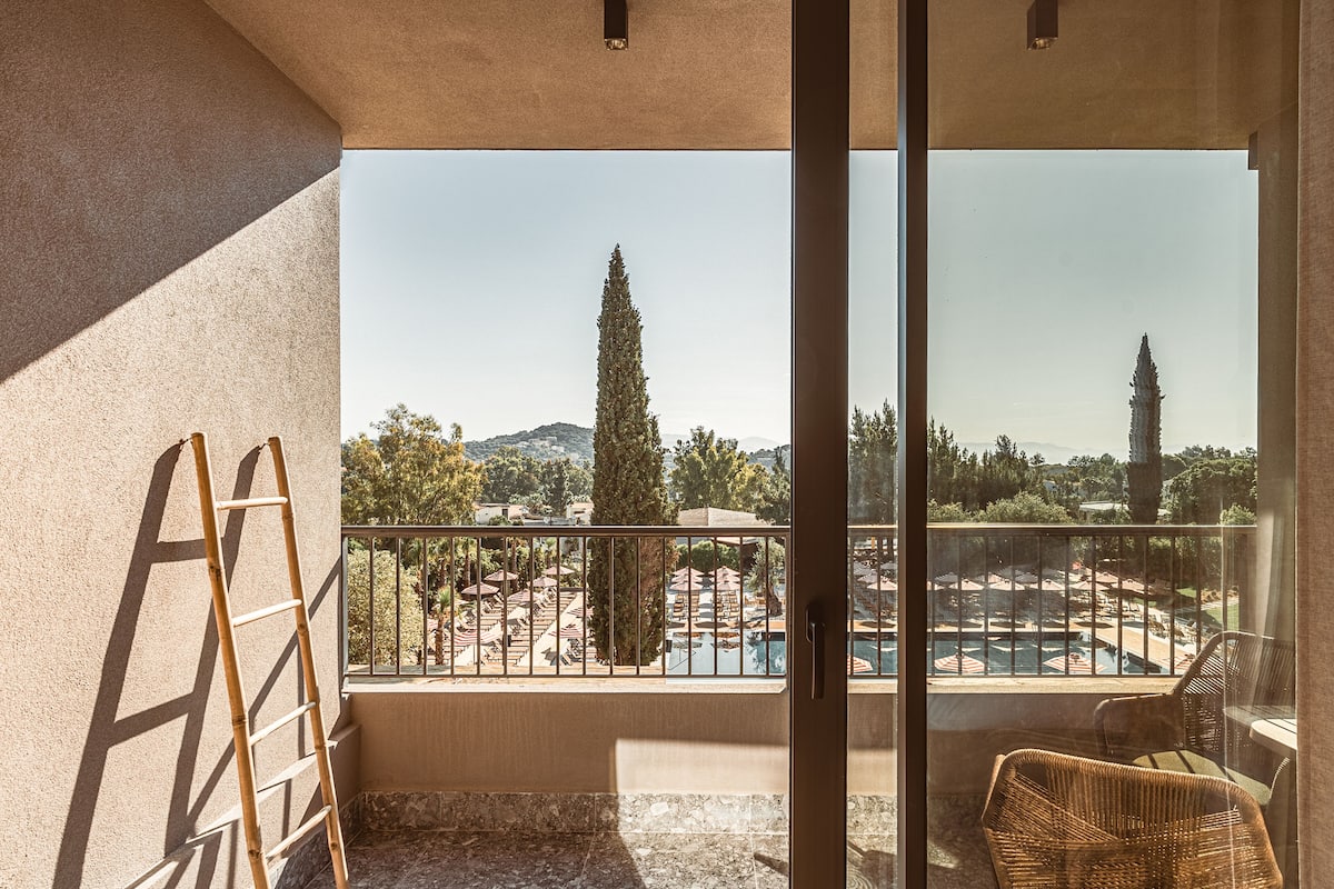 A balcony with a view of a pool and trees, a wicker chair, and a wooden ladder.