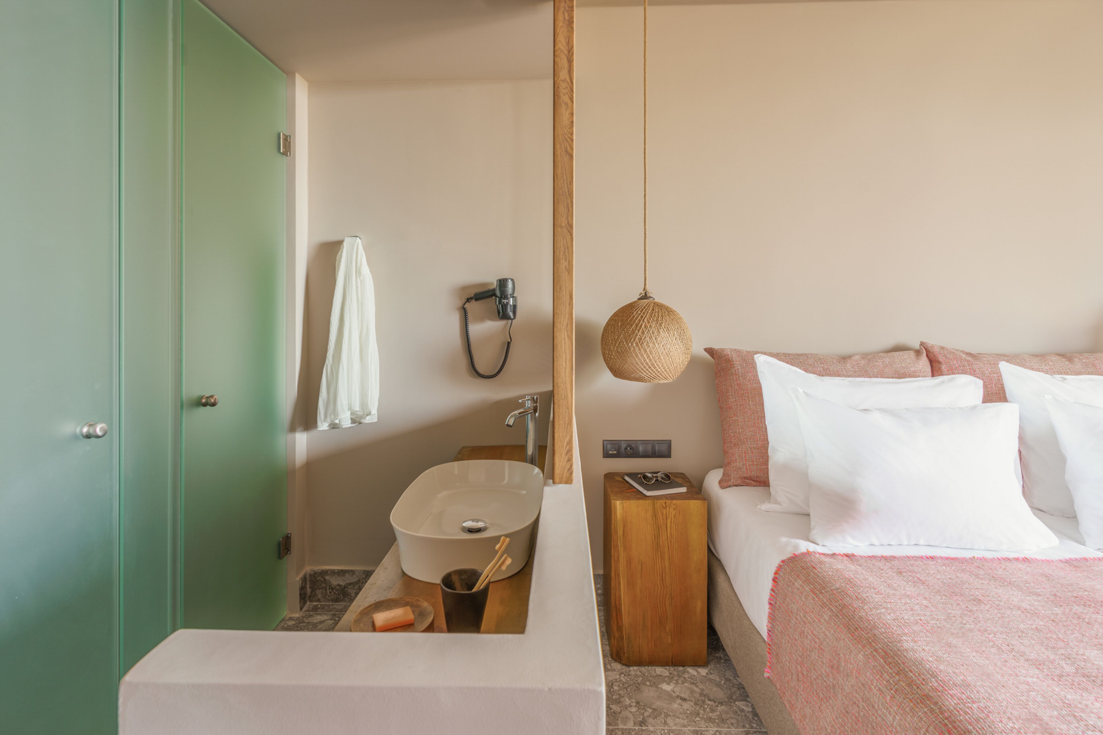 Habitación de hotel con un armario verde, un lavabo blanco, una mesita de noche de madera, una lámpara colgante y una cama con ropa de cama blanca y rosa.