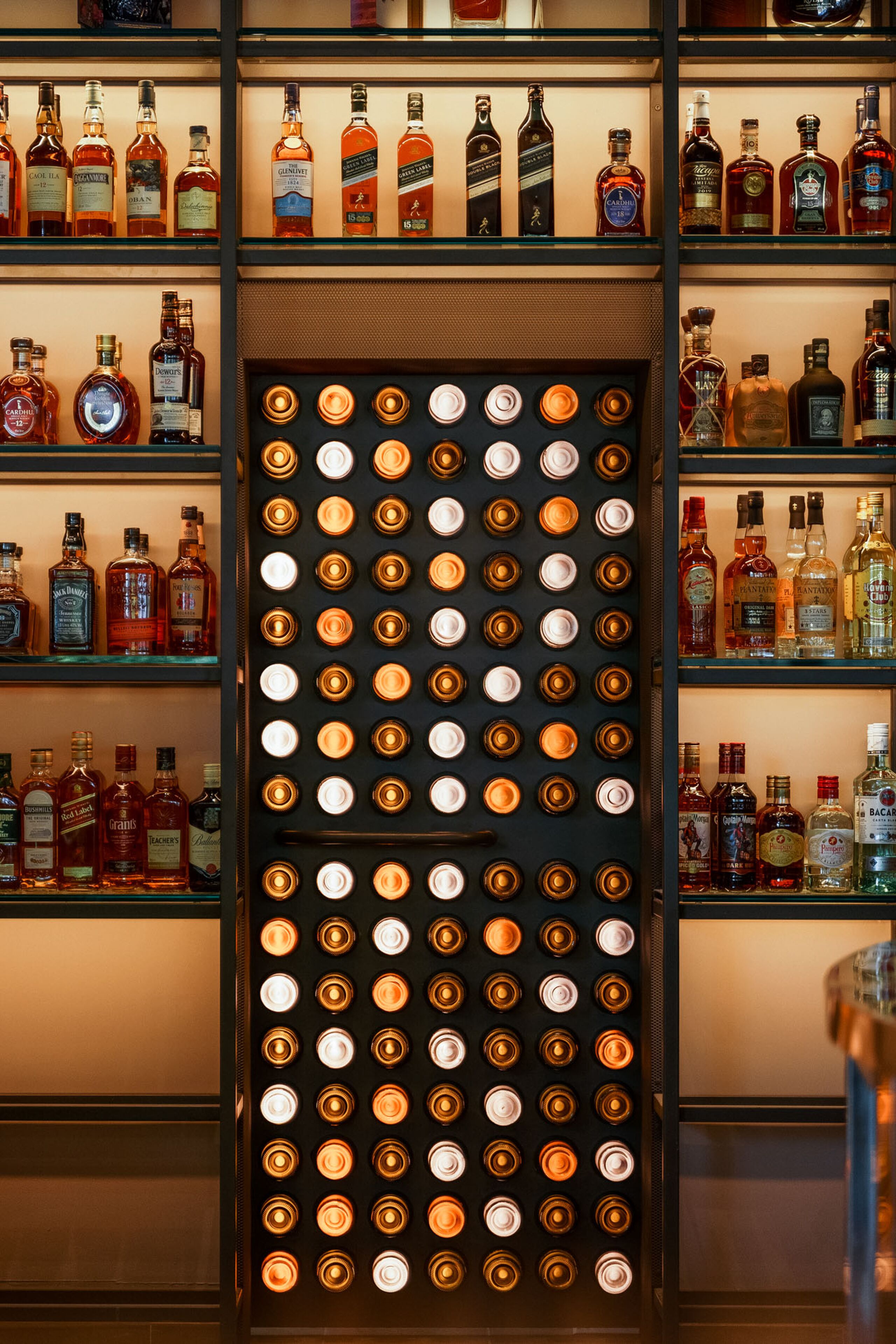 Una licorería con estantes llenos de botellas diversas y una pared decorada con tapones de botella iluminados.
