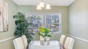 Une salle à manger avec une table en bois, des chaises blanches, une plante en pot et une fenêtre avec des stores.