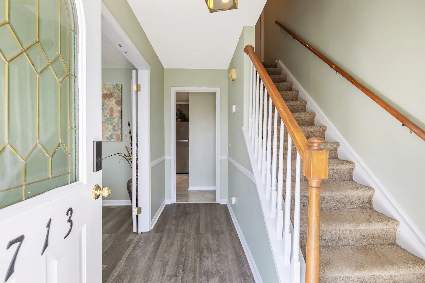 Un couloir avec un escalier en bois, une porte portant une plaque avec un numéro, et un luminaire au plafond.