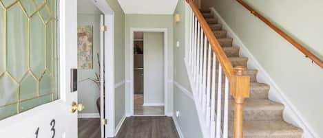 Un couloir avec un escalier en bois, une porte portant une plaque avec un numéro, et un luminaire au plafond.