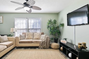 Una sala de estar con un sofá color beige, un ventilador de techo, un televisor y un ventanal con persianas.