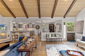 A spacious living room with a wooden ceiling, a dining area with a long table, and a kitchen in the background. - Pacific Serenade: Brand New Remodel (Carmel-by-the-Sea)