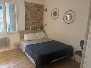 A bedroom with a brick wall, a bed with striped bedding, two wall-mounted mirrors, and a window with blinds. - Charming, renovated studio apartment in Toulouse, in a beautiful location (Toulouse)