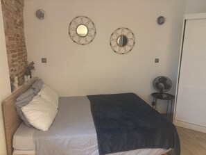 A bedroom with a bed, two wall-mounted mirrors, a small table with a fan, and a brick wall. - Charming, renovated studio apartment in Toulouse, in a beautiful location (Toulouse)