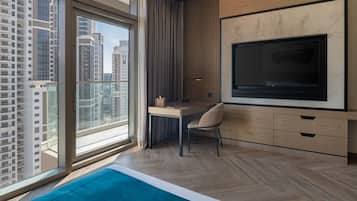 A modern hotel room with a large window, a desk with a chair, a flat-screen TV, and a bed with a blue accent.