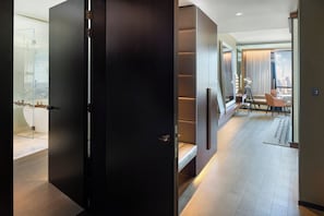 A modern hotel room with a dark wooden door, a glass shower, a white bathtub, a seating area with a table and chairs, and a city view through large windows.