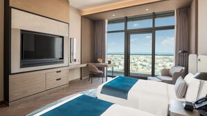 Habitación de hotel con un televisor grande, un escritorio con una silla, un sofá y una cama con una manta azul.