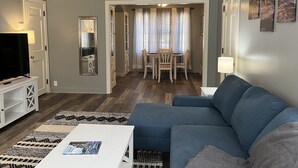 A living room with a blue sofa, a white coffee table, and a TV on a stand. There is a dining area with a table and chairs. - Historic Mc Kennan Park Apt 2 (1 bed 1 bath) - KP220APT2 (Sioux Falls)