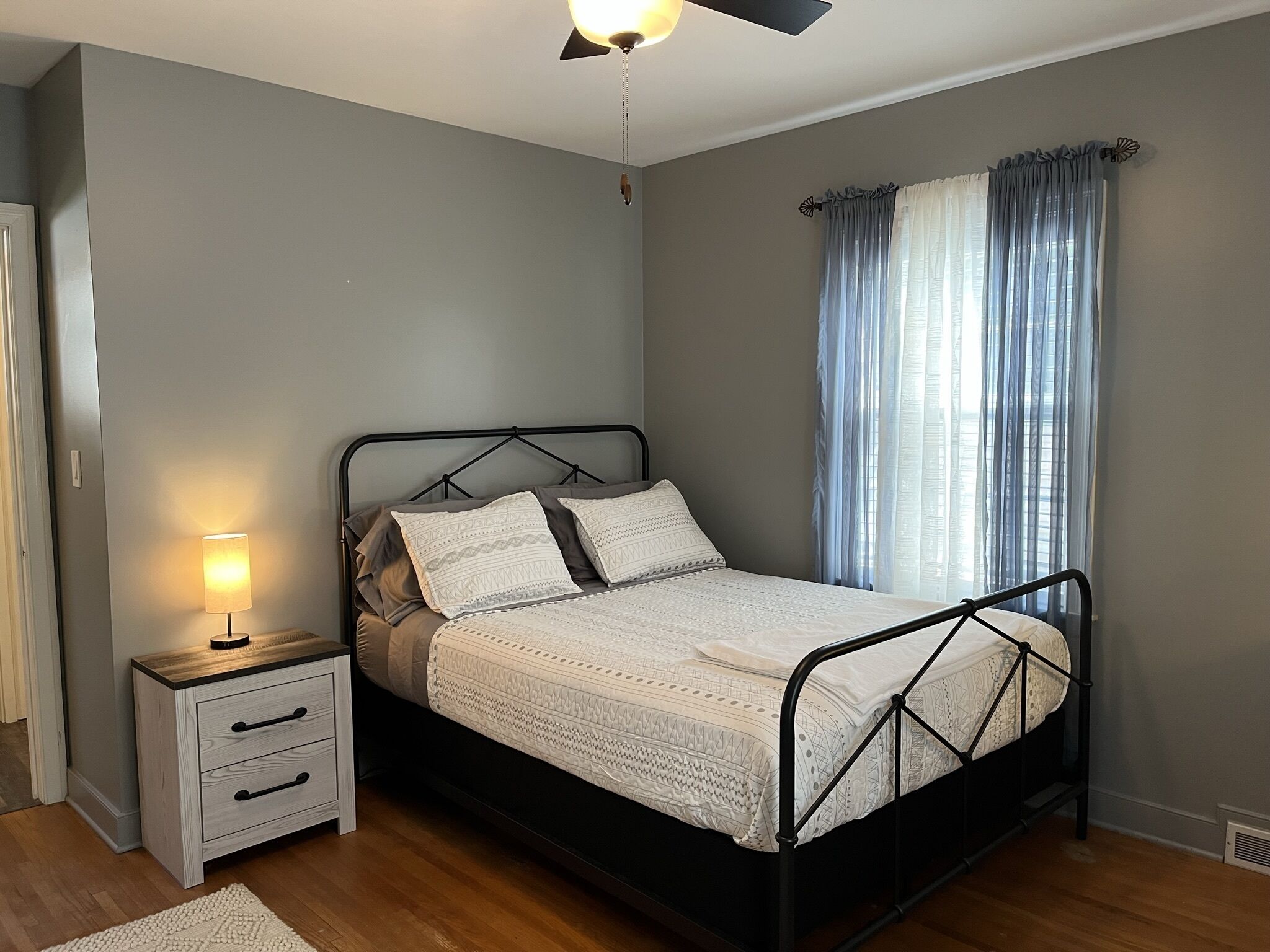 Ein Schlafzimmer mit Bett, Nachttisch, Lampe und Fenster mit Vorhängen.