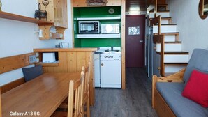 A compact kitchen with a microwave, a small refrigerator, and a sink. There is a wooden bench and a sofa with a red cushion. A staircase leads to an upper level.