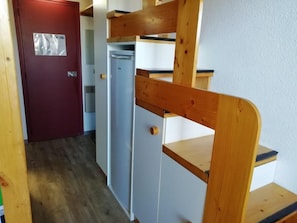 A compact kitchen with a refrigerator, wooden shelving, and a red door.