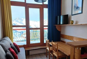 A room with a sofa, wooden table, chairs, a TV, and a view of snow-capped mountains through large windows.