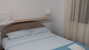 En säng för en person med en sänggavel, två kuddar, ett nattduksbord med en lampa och ett fönster med tunna, genomskinliga gardiner.