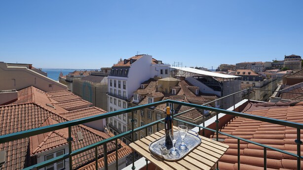 Eine Terrasse mit Tisch für zwei Personen, mit Blick auf eine Stadtlandschaft mit terrakottafarbenen Dächern und blauem Himmel.