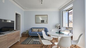 A modern living room with a blue sofa, white chairs, a dining table, and a large mirror on the wall.