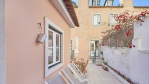 A narrow outdoor corridor with a white tiled floor, a table with chairs, and a white wall with a window and a metal bucket.