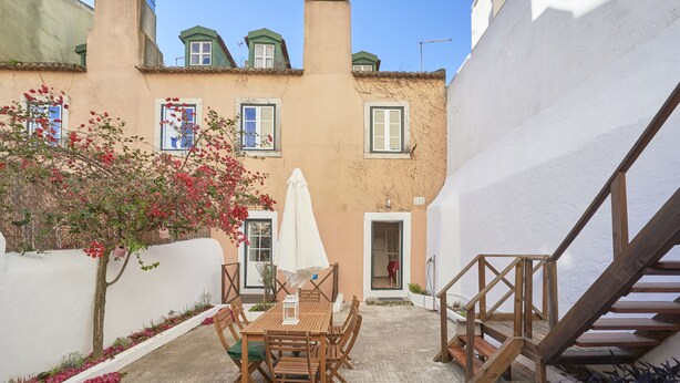 Une cour avec une table et des chaises en bois, un mur blanc, un escalier et un arbre aux fleurs rouges.