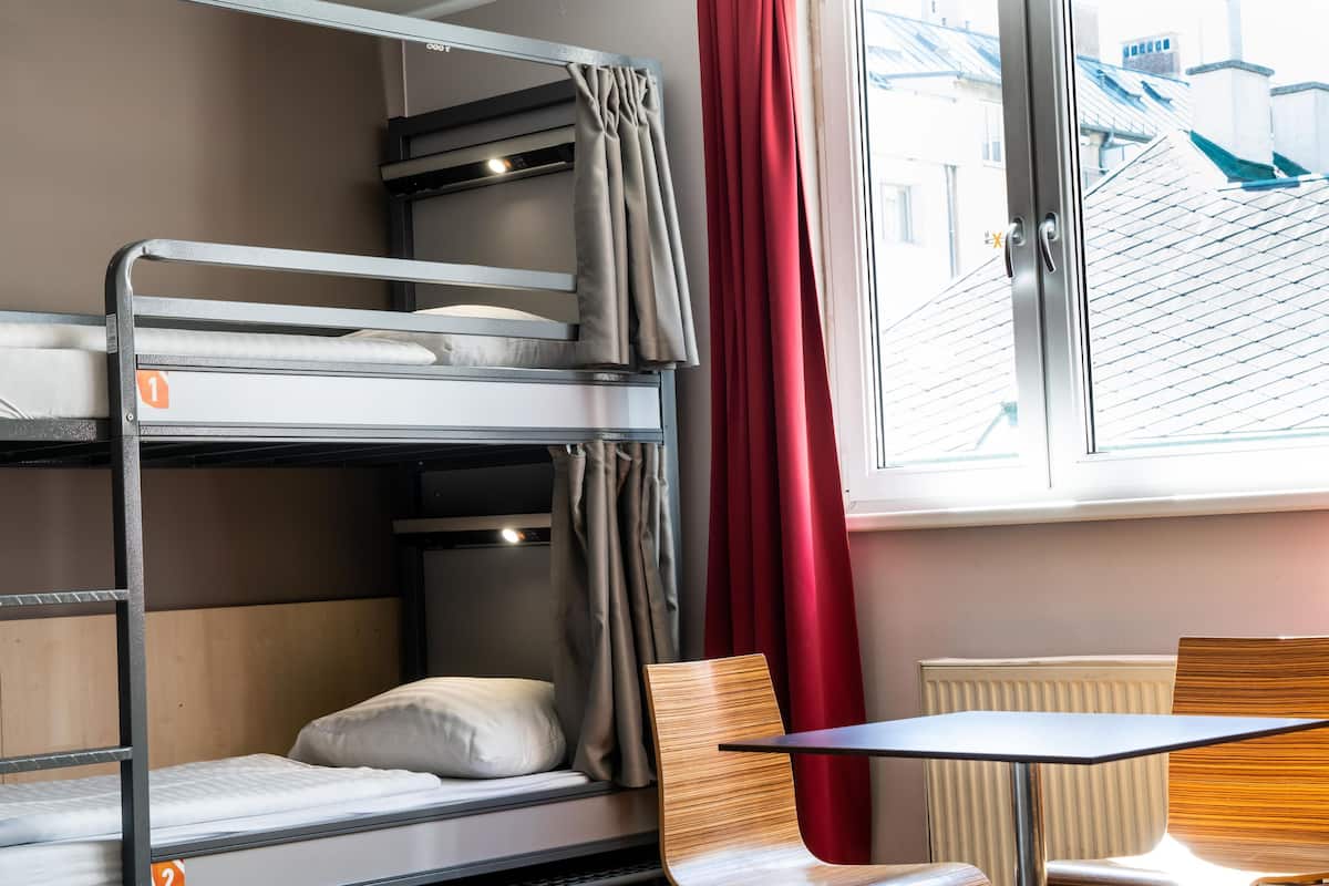 A bunk bed with a window, a table, and a chair in a room with a radiator and curtains.