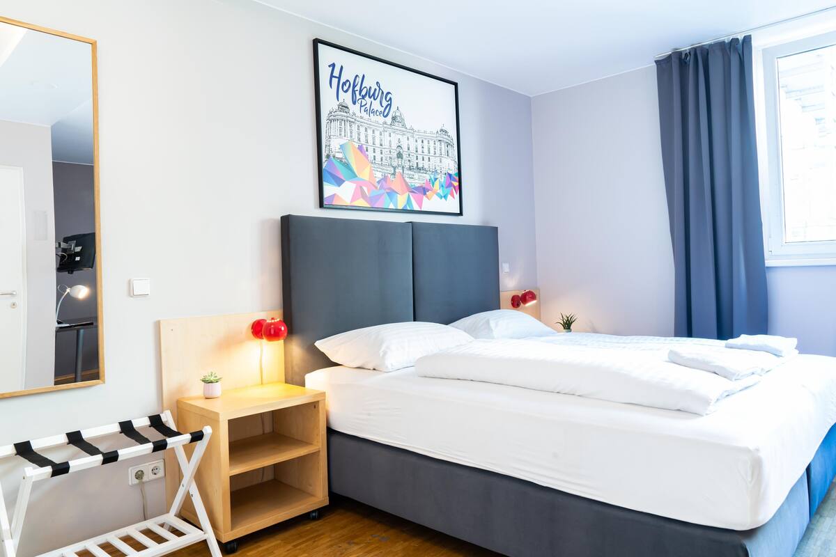 A hotel room with a bed, a nightstand, a mirror, a framed picture of a building, and a window with curtains.