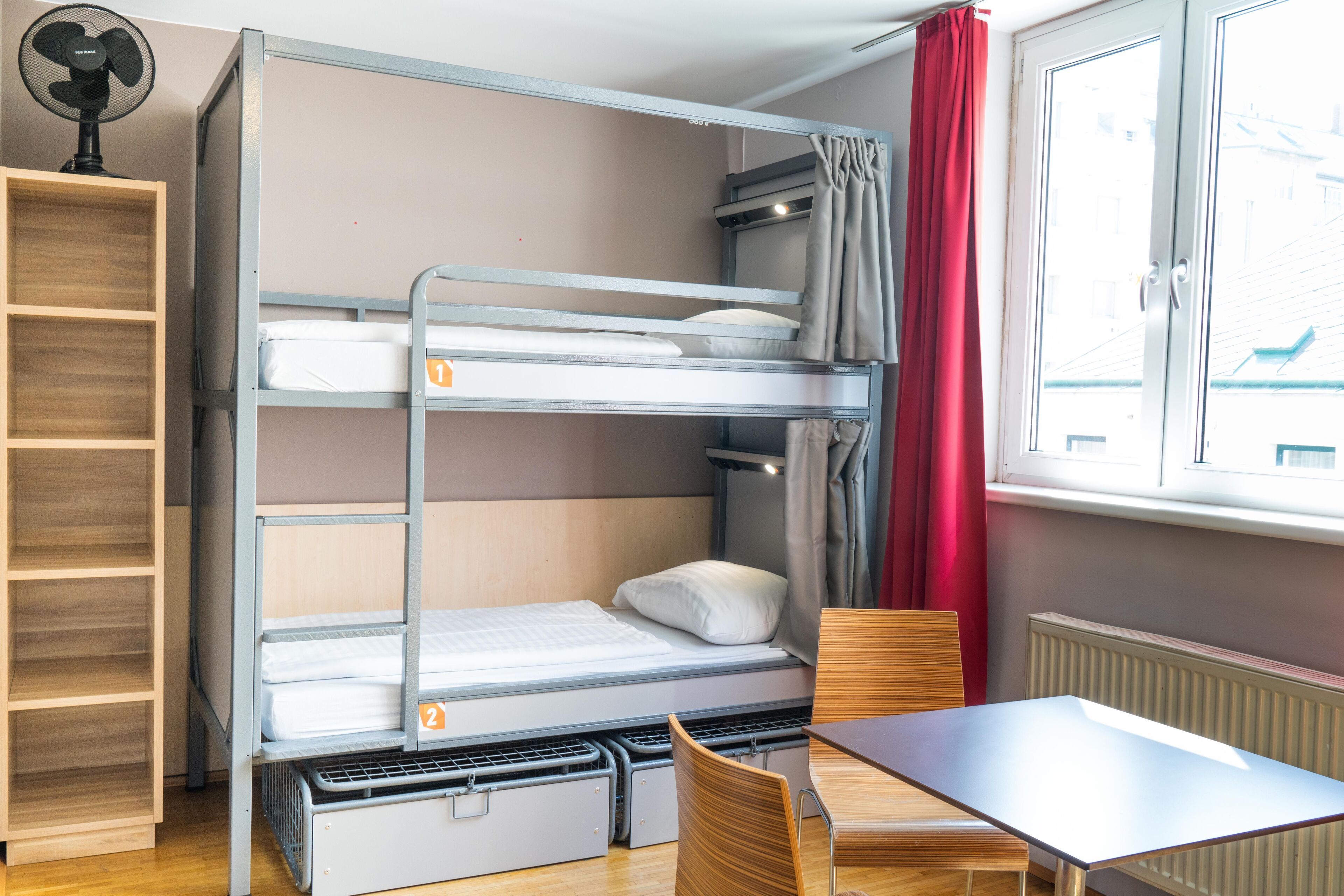 Uma cama beliche com dois níveis, uma estante de madeira, uma mesa com cadeiras e uma janela com cortinas.
