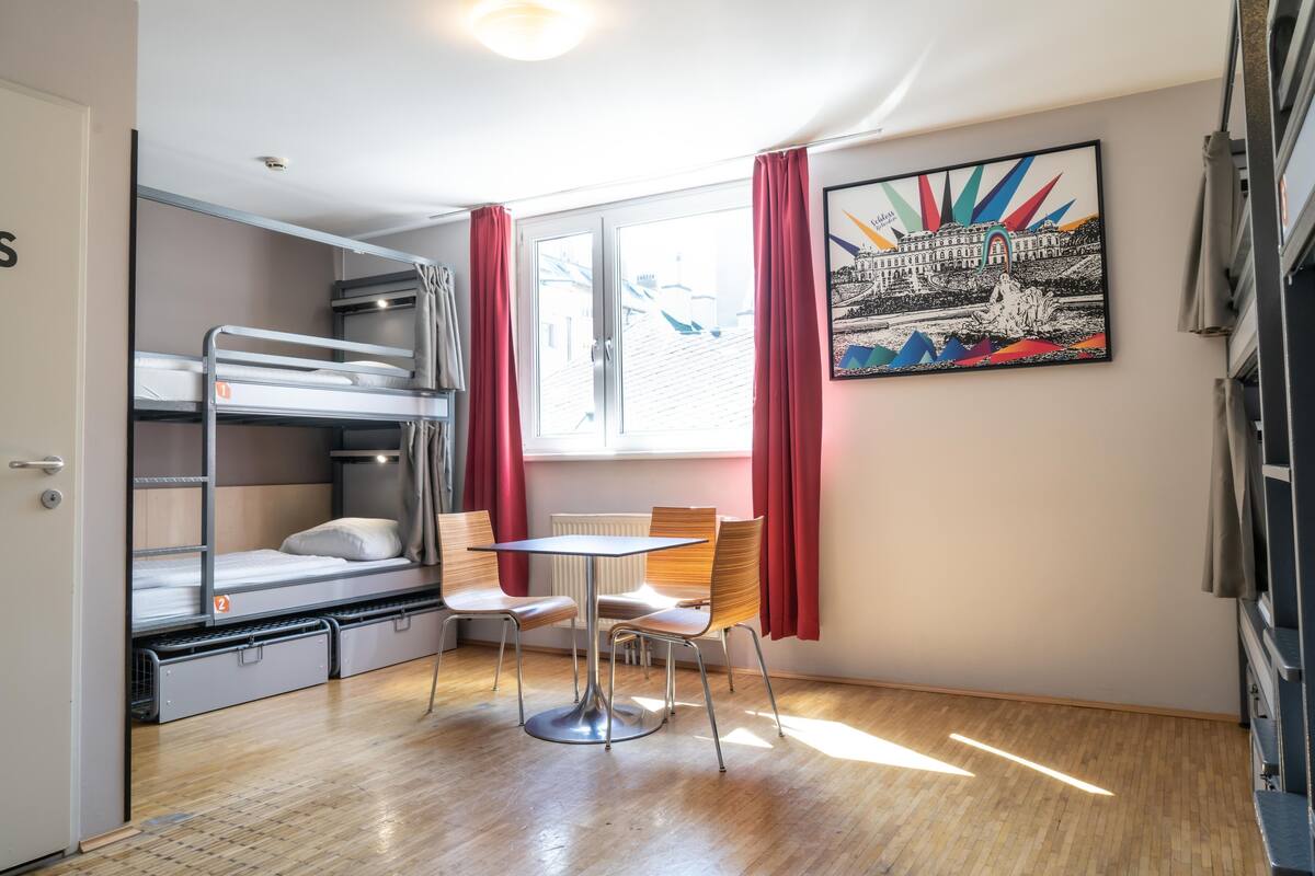 A dormitory room with bunk beds, a table, chairs, a radiator, and a window with curtains.