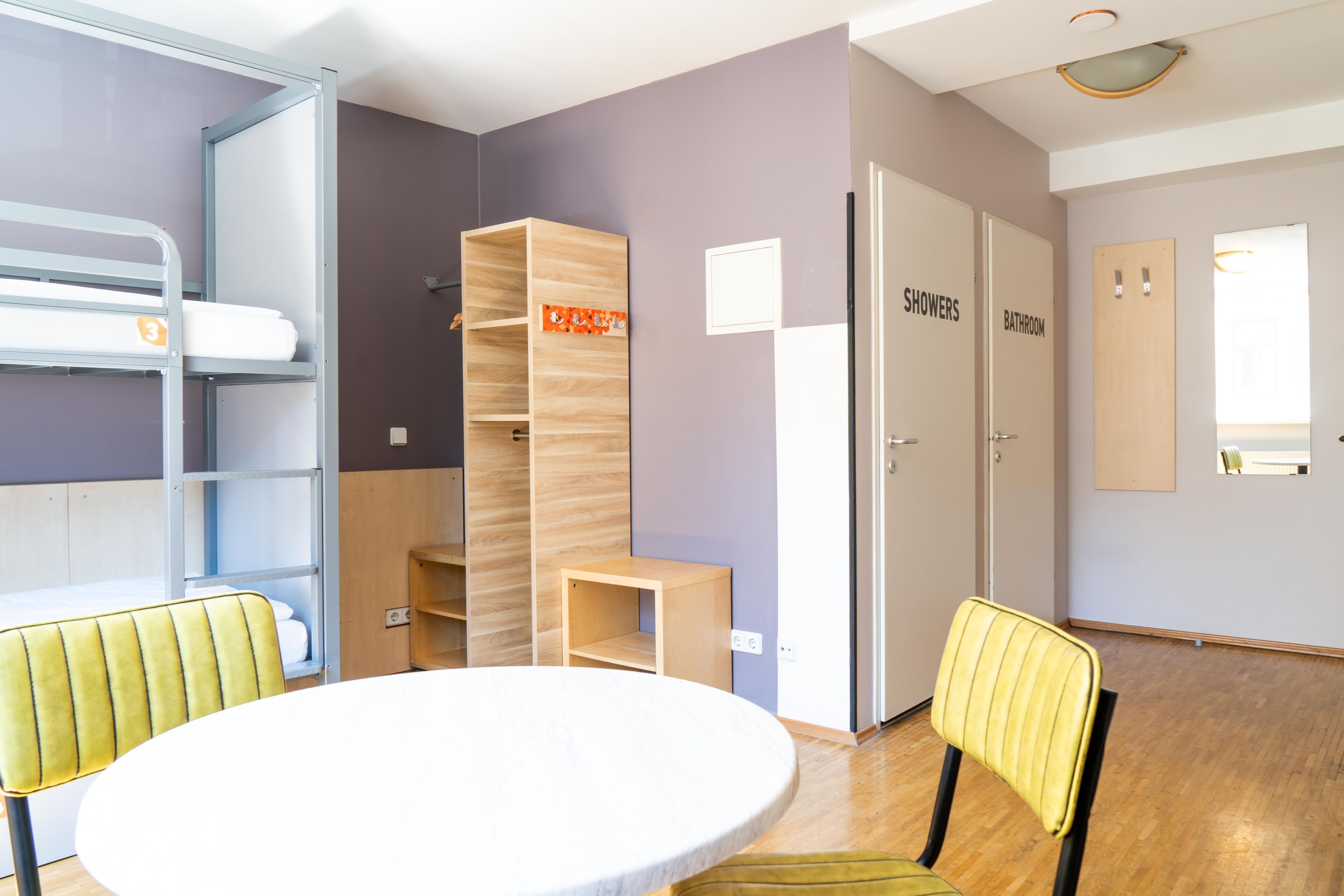 A room with bunk beds, a round table, chairs, and two doors labeled "Showers" and "Bathroom".
