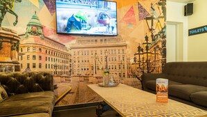 A hotel room with a large mural of a cityscape, a television, a sofa, a coffee table, and a sign indicating the location of the toilets. - St Christopher's Vienna - Hostel (Vienna)