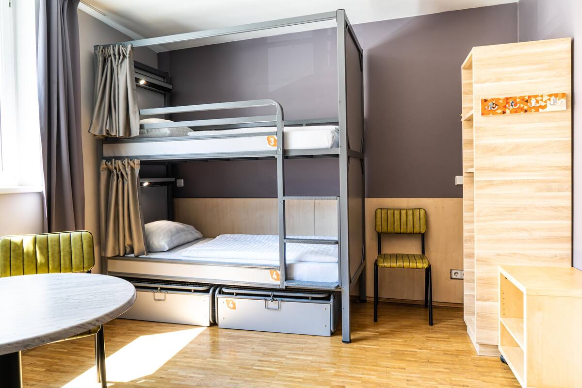 A bunk bed with a desk and chair, a small table, and a wooden cabinet in a room with wooden flooring and a window with curtains.