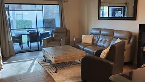 A spacious living room with a large window, a ceiling fan, a leather sofa, a coffee table, and a TV. - Gulf Gate - Quaint and fresh 1st floor condo w/covered parking (Sarasota)