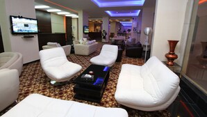 A modern lounge area with white leather sofas, a flat-screen TV mounted on the wall, and a patterned carpet.