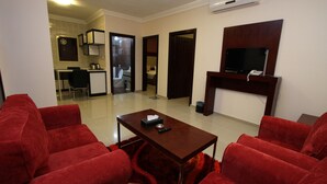 A living room with red sofas, a wooden coffee table, a TV mounted on the wall, and a kitchen area in the background.