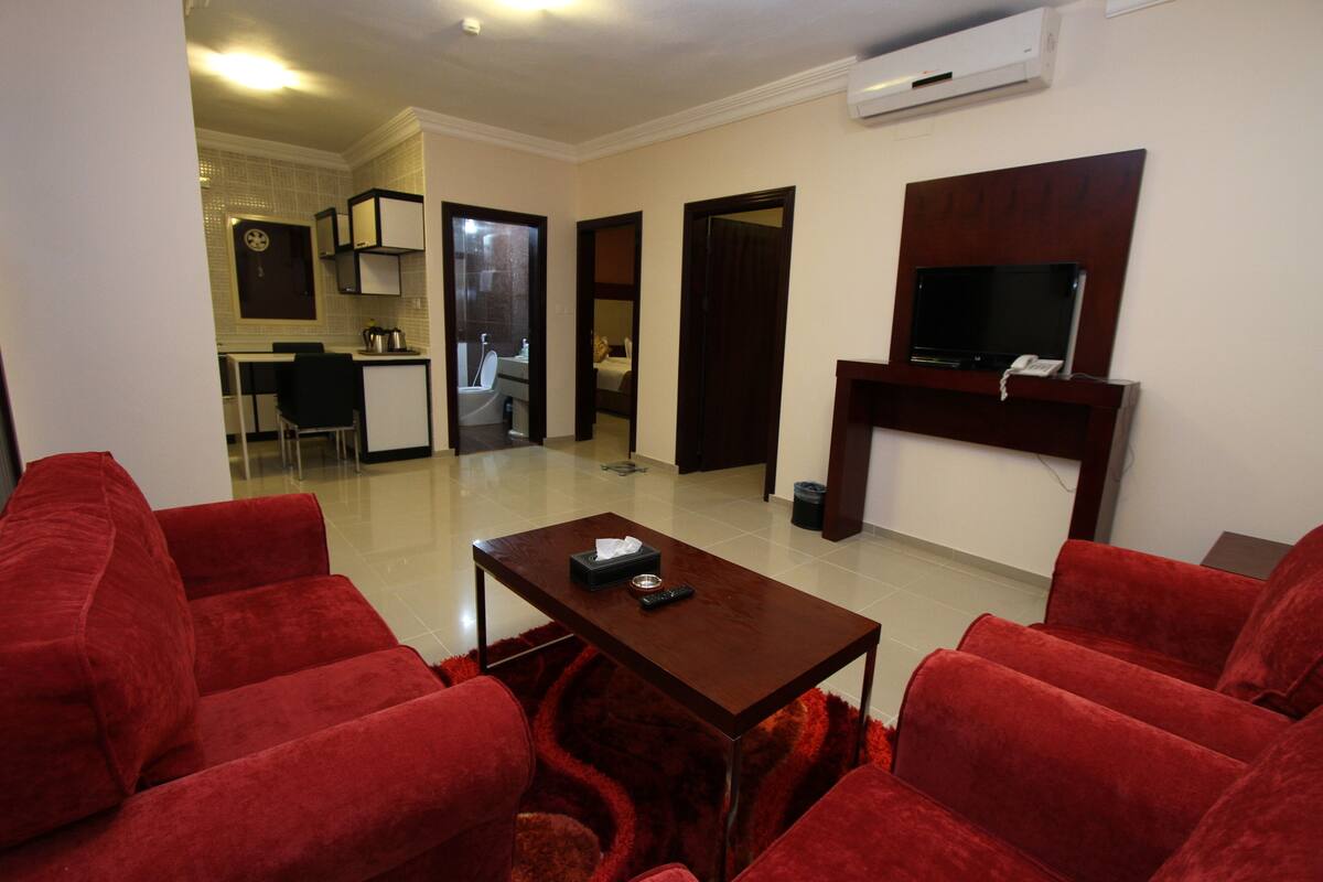 A living room with red sofas, a wooden coffee table, a TV mounted on the wall, and a kitchen area in the background.