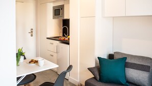 A compact kitchenette with a microwave, a small refrigerator, and a countertop with fruits and a toaster. There is a dining area with a white table, two chairs, and a vase with flowers. A grey sofa with teal cushions is visible in the foreground.