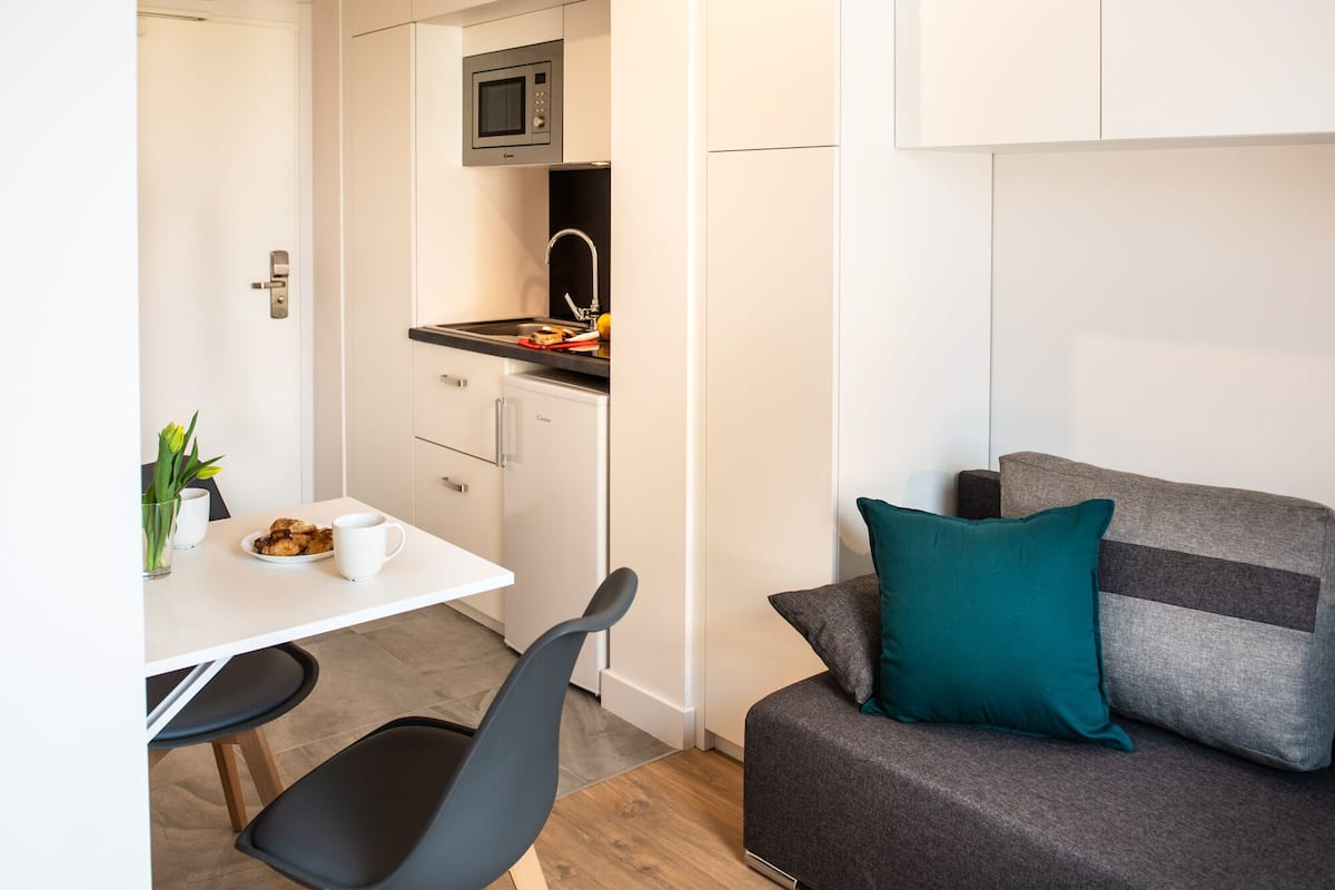 Una kitchenette compacta con microondas, un refrigerador pequeño y una encimera con frutas y un tostador. Hay una zona de comedor con una mesa blanca, dos sillas y un jarrón con flores. Un sofá gris con cojines color turquesa es visible en primer plano.