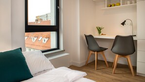 A bedroom with a bed, a window with a view of buildings, a desk with two chairs, and a shelf with apples and cups.