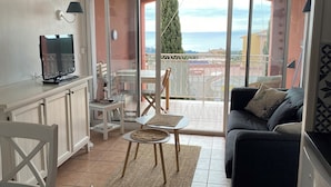 Un salon avec un canapé, une table basse et un téléviseur posé sur un meuble. Il y a un balcon avec vue sur la mer.