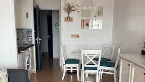 Un salon moderne avec un canapé, une table basse et un coin repas comprenant des chaises et une table. On aperçoit un lave-linge au fond de la pièce, et le sol est carrelé.