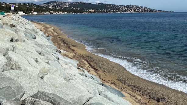 Une plage rocheuse offrant une vue dégagée sur la mer et sur une colline lointaine où se trouvent des bâtiments.